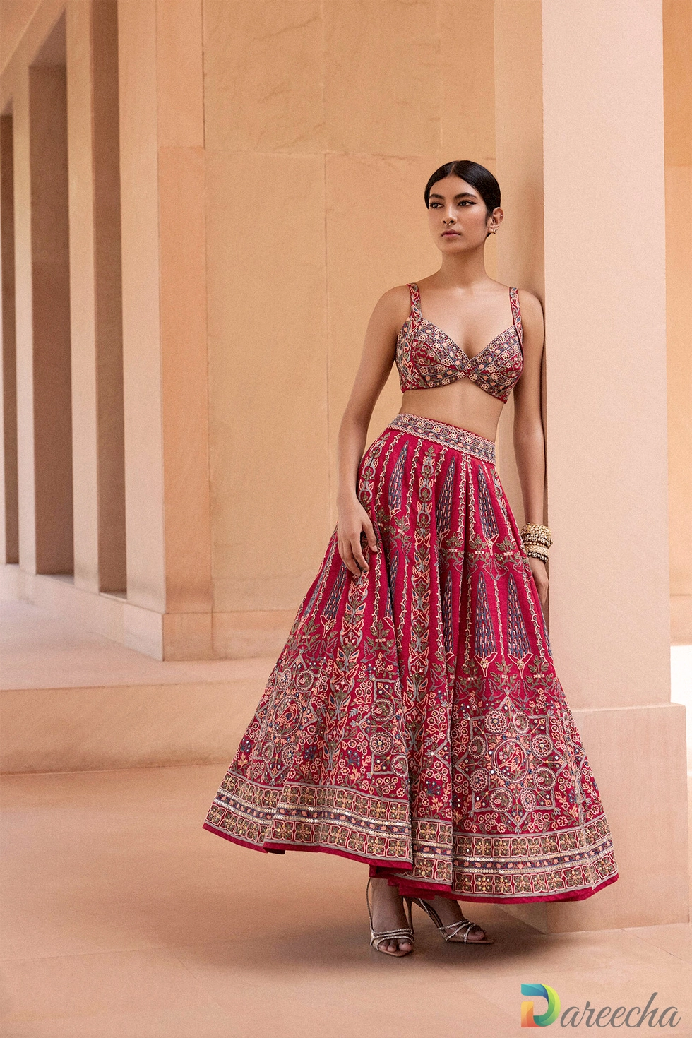 Model wearing vintage red matka silk lehenga with thread and mirror hand embroidery standing between beige stone arches