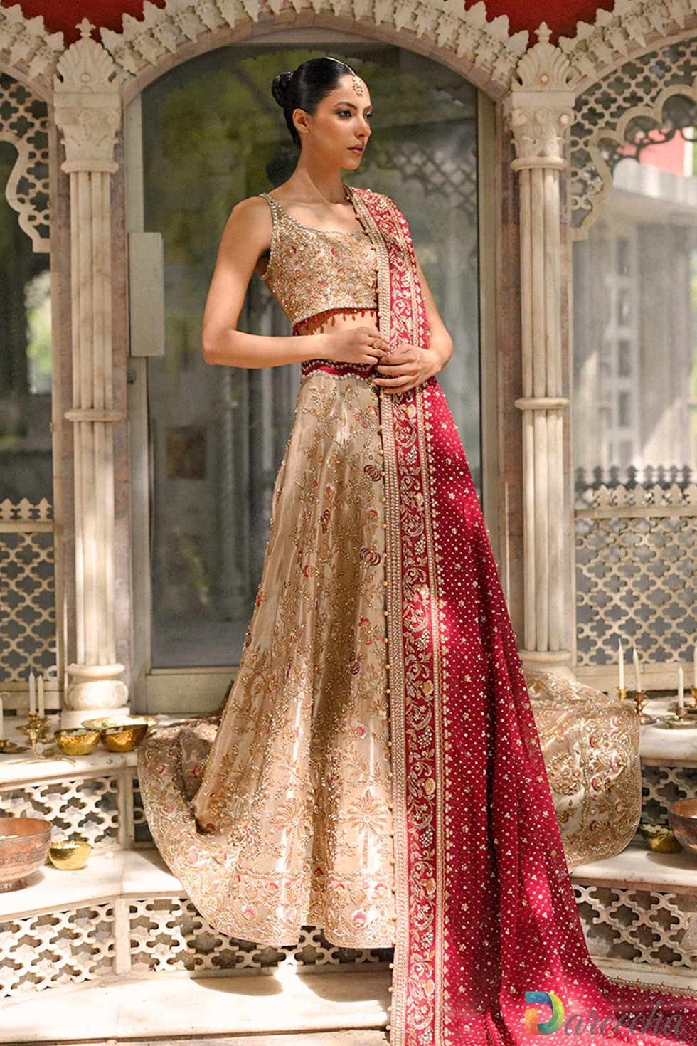 Full length view of model in gold tissue lehenga choli set with crimson cotton net dupatta standing under arches