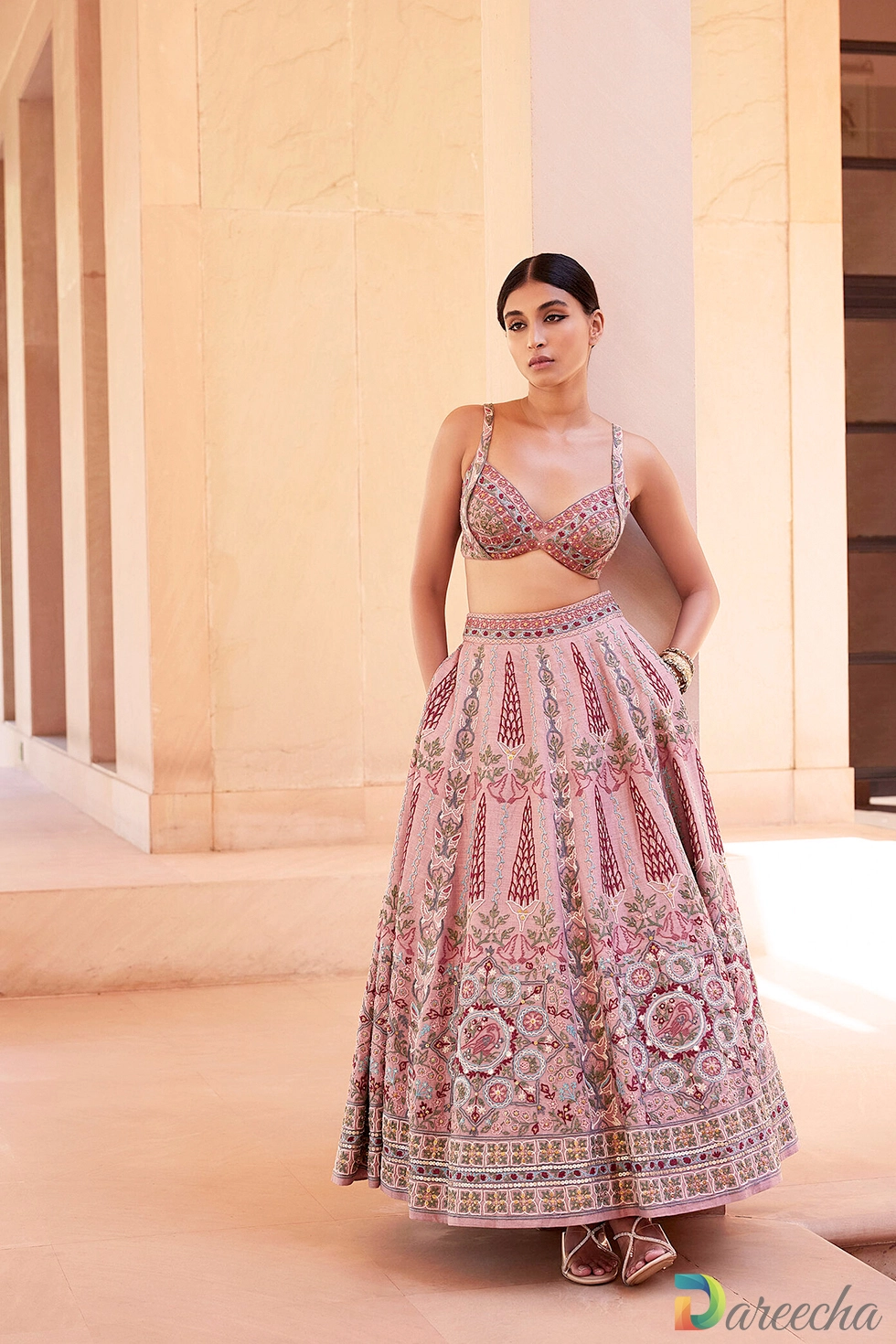 Model wearing dusty rose matka silk embroidered lehenga skirt and sweetheart bustier top with hands in pockets standing between sandstone pillars