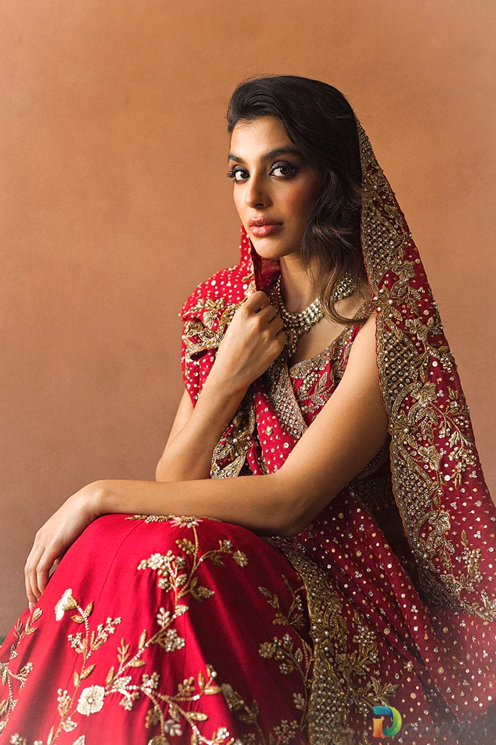Seated profile shot of model in candy red raw silk bridal lehenga wearing dupatta draped over h Seated profile shot of model in candy red raw silk bridal lehenga wearing dupatta draped over h
