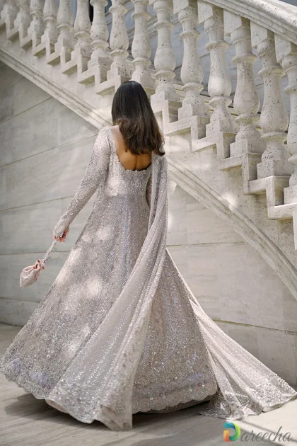 Back view of model walking up marble stairs showing backless scoop neckline of champagne pink peshwas flowing dupatta train and matching hand embroidered pouch