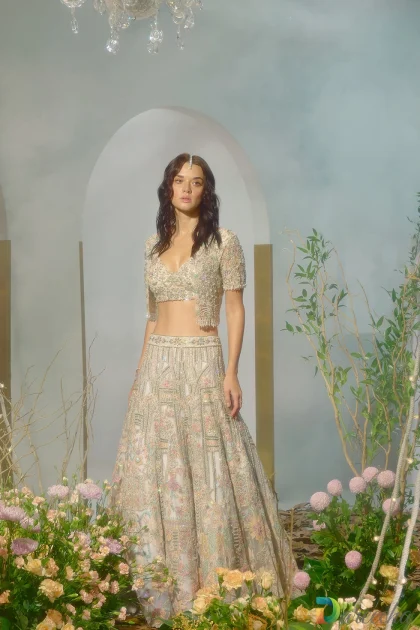 Full length standing portrait of white lehenga jacket set with flared tulle lehenga and lush floral background