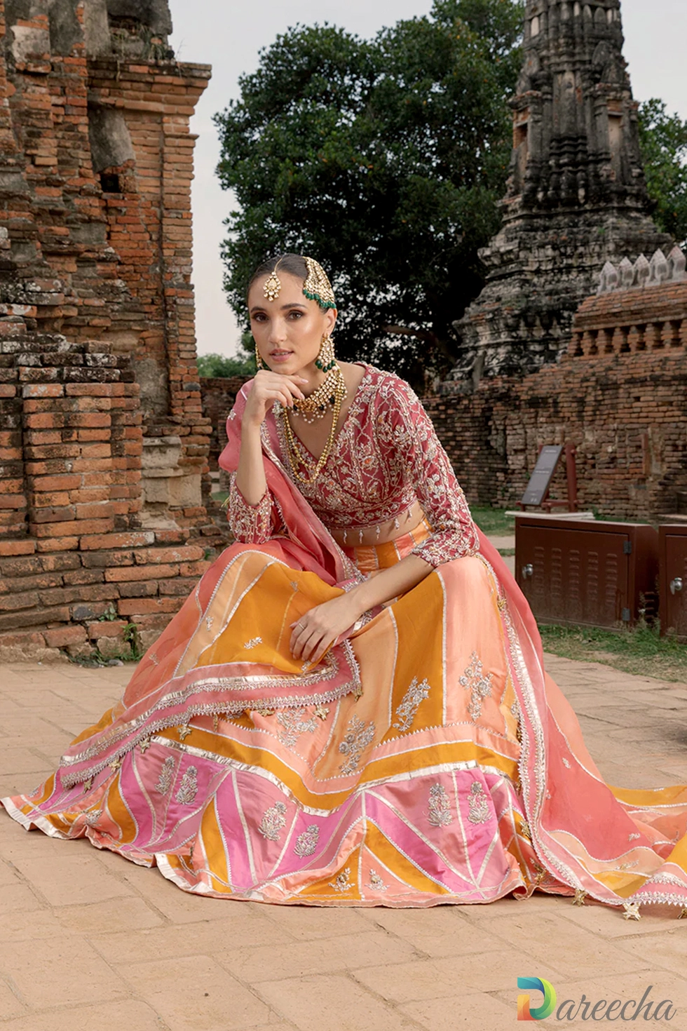 Model sitting against brick backdrop wearing multi color chatta pati lehenga with rust sheesha silk blouse