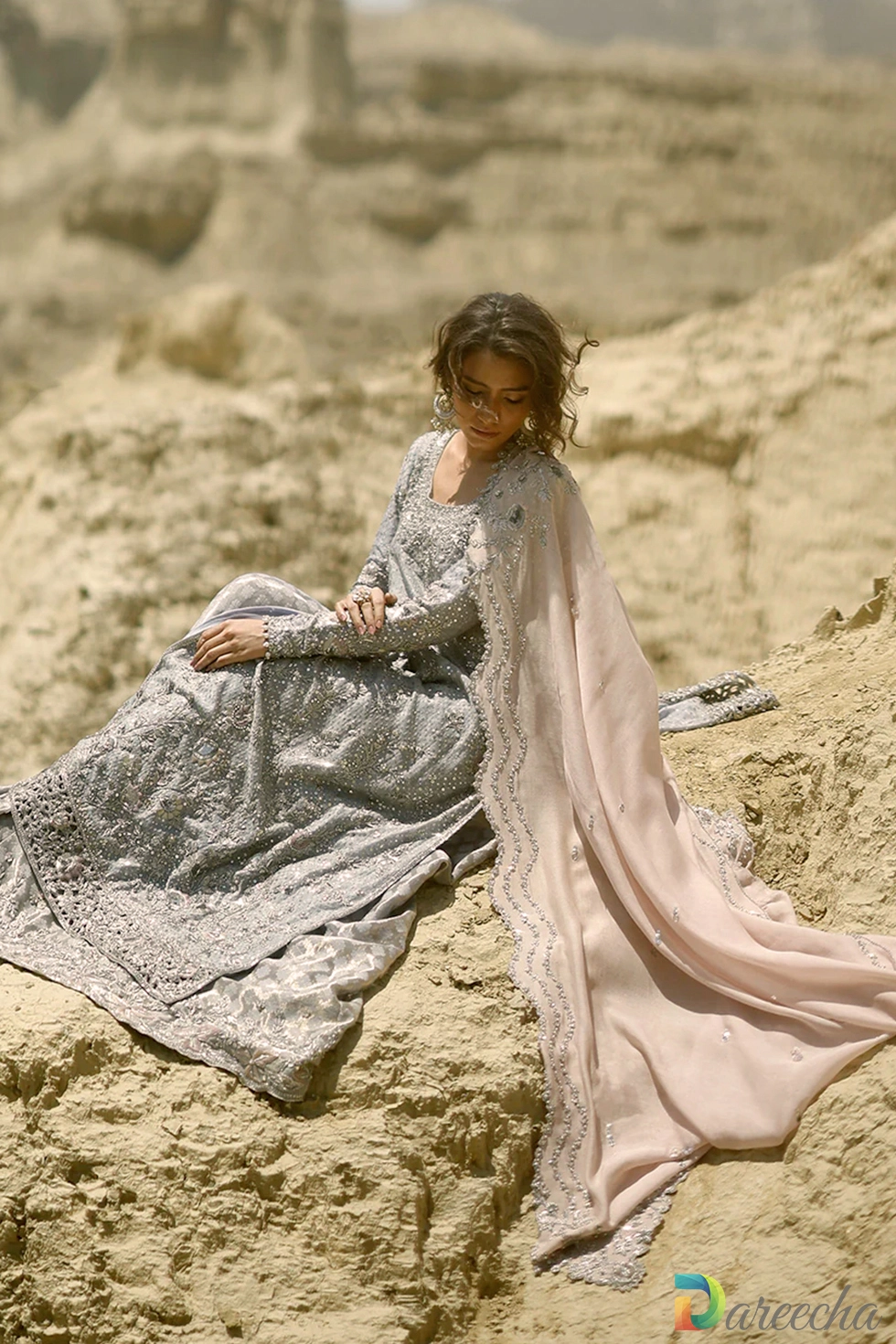 Seated pose on rocks displaying sequined diamond trellis on steel blue long shirt with lehenga and blush dupatta