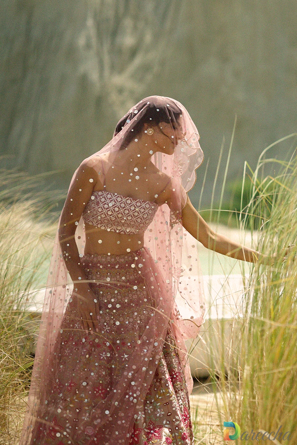 Seated pose in high grass showing pink tulle angrakha lehenga dupatta draped as a sheer veil
