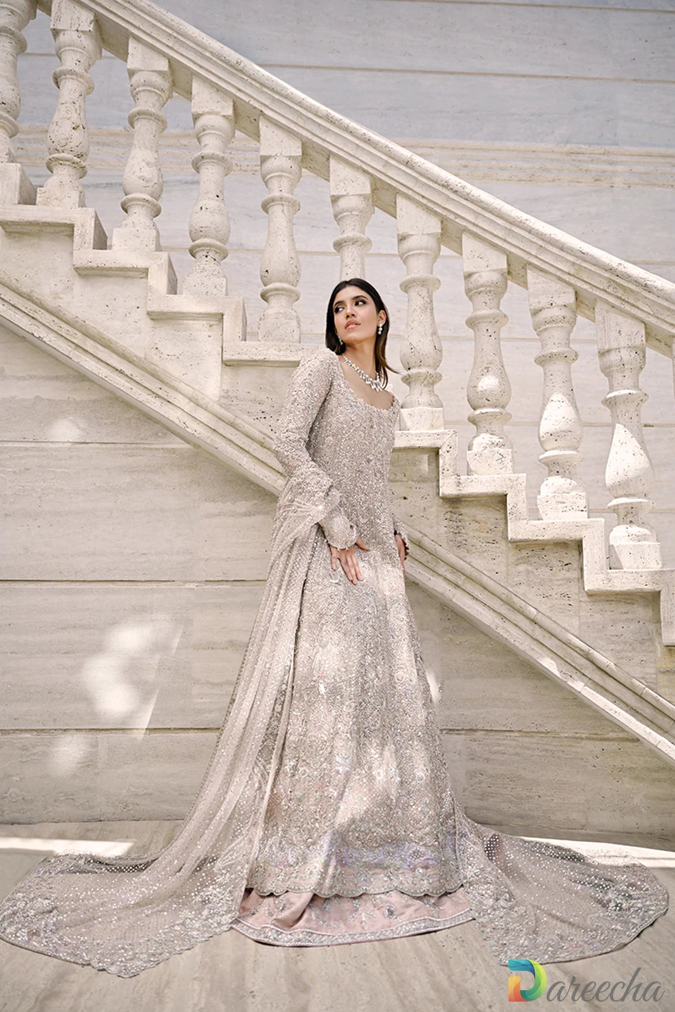 Full length front view of model posing on marble staircase displaying flared peshwas skirt over layered brocade lehenga