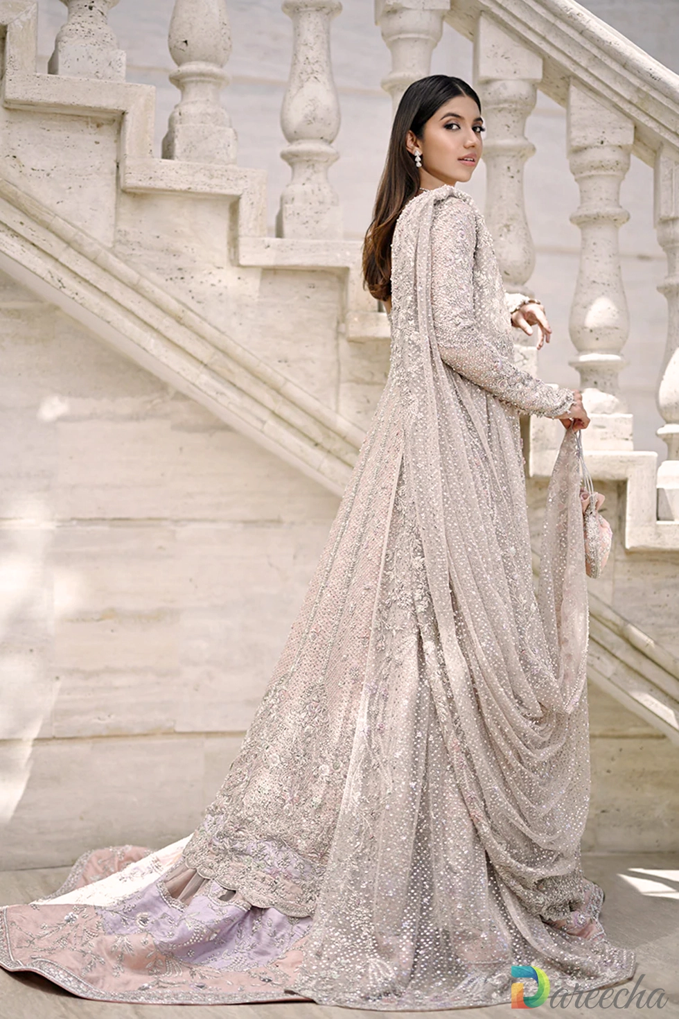 Side profile view of model looking back on marble staircase showing draped tulle dupatta embroidered sleeves layered skirt hem and matching pouch