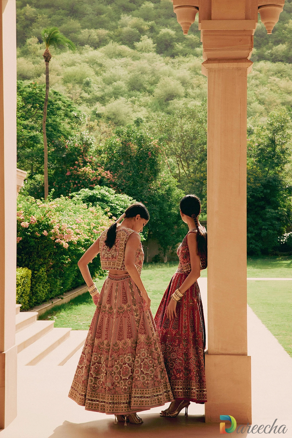 Back view of model wearing dusty rose embroidered matka silk lehenga set standing beside model in red lehenga in outdoor colonnade