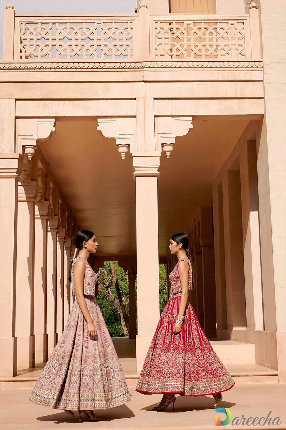 Two models standing in an arcade wearing handcrafted red and dusty pink embroidered matka silk lehenga sets