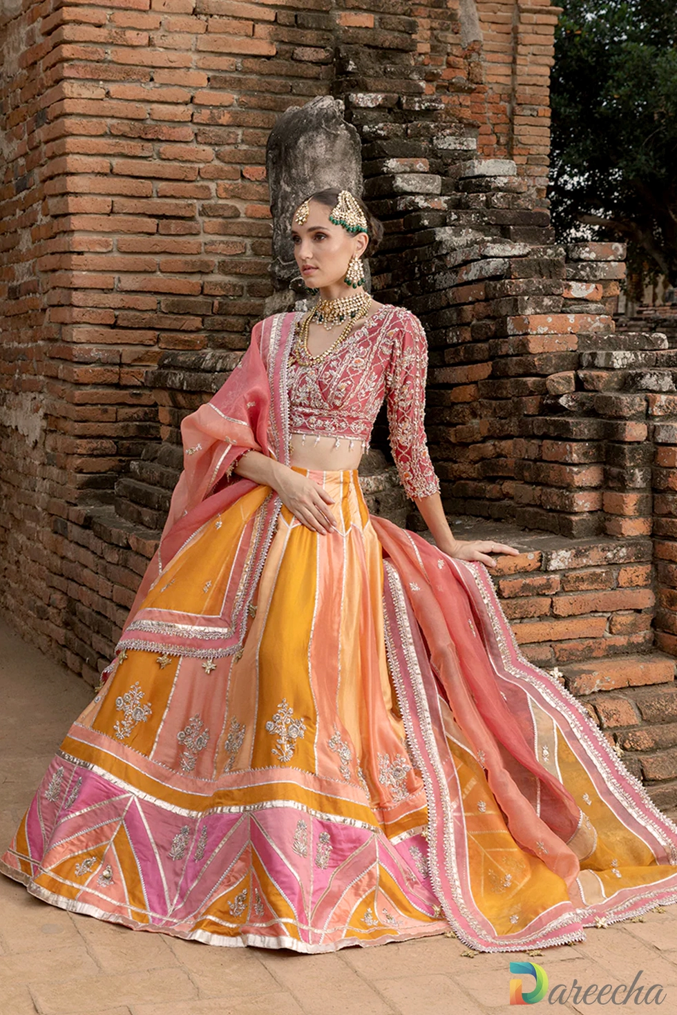 Seated pose against brick wall highlighting zardozi embroidery on rust blouse and multi color chatta pati lehenga
