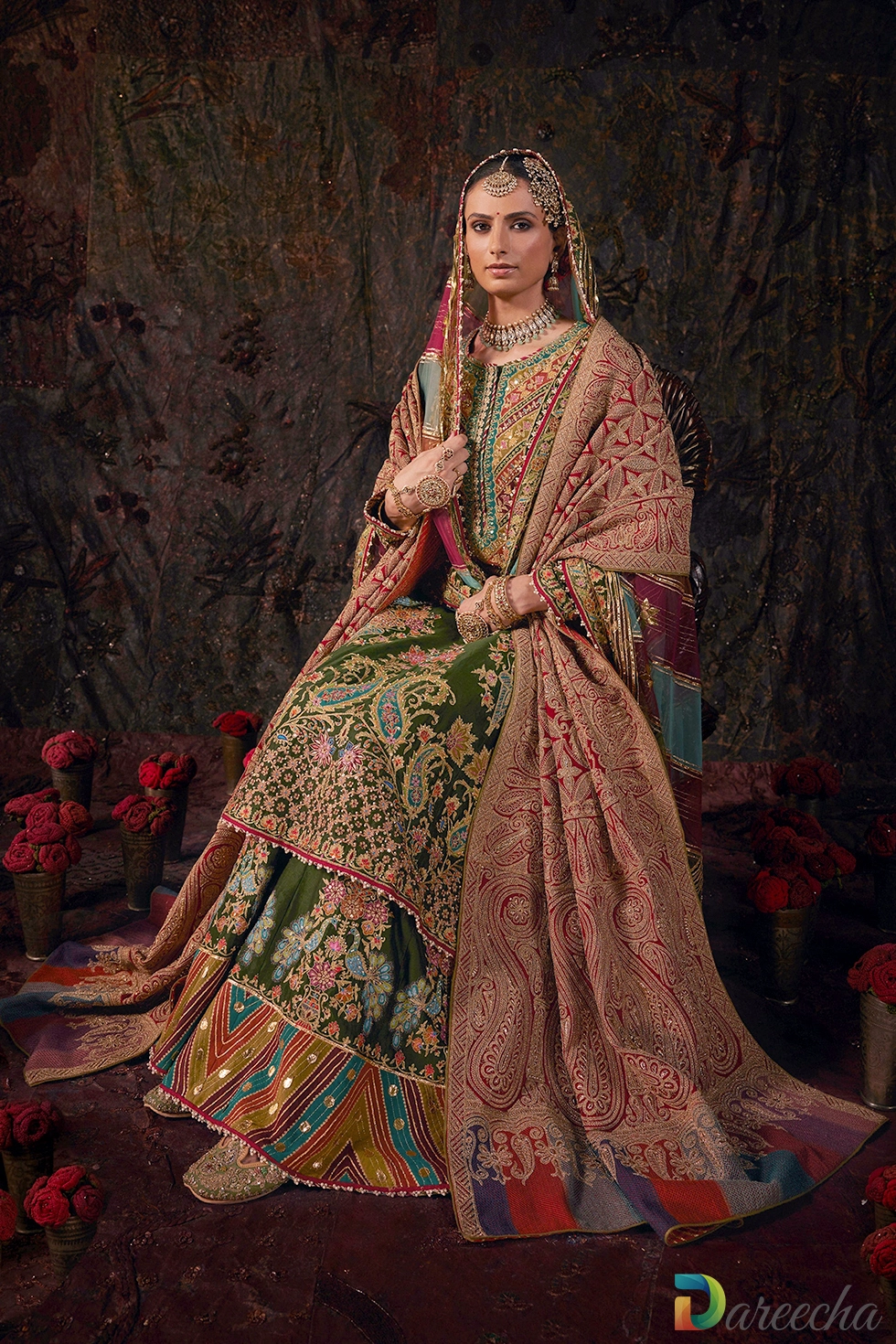 Model seated wearing forest green silk sharara with embroidered dushala, multi hued head dupatta, and peacock motifs