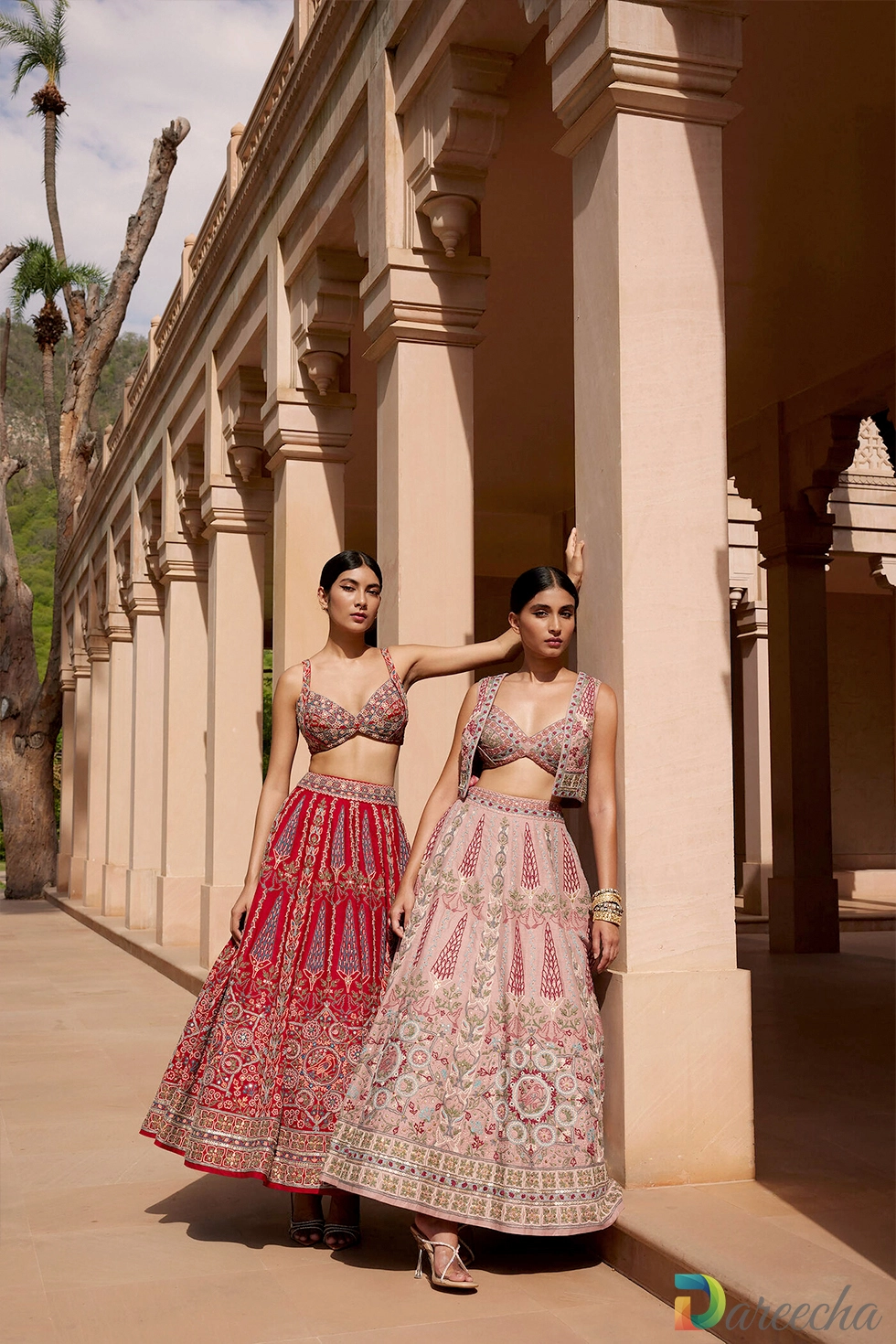 Two models standing in an arcade wearing handcrafted red and dusty pink embroidered matka silk lehenga sets