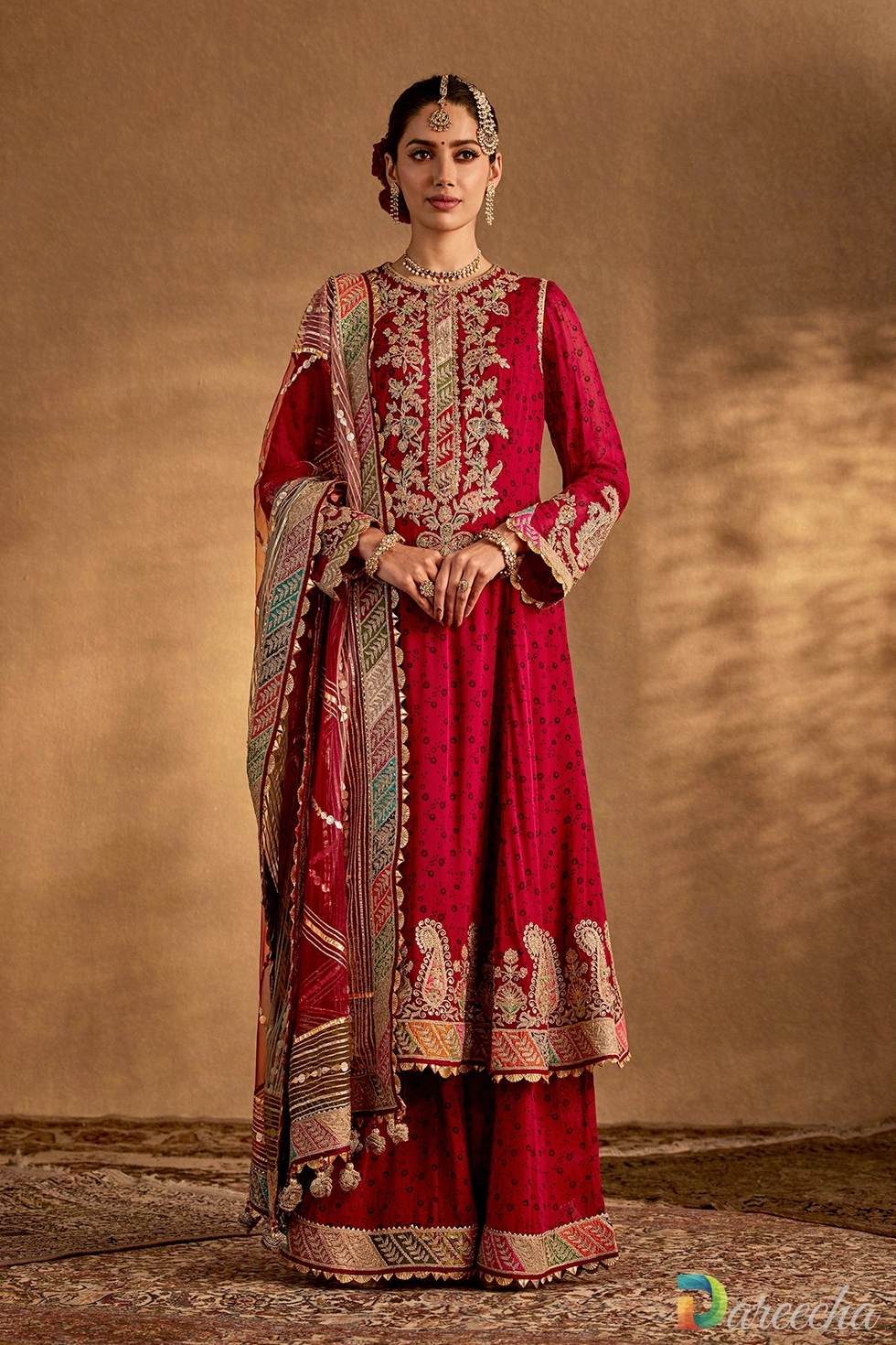 Model wearing an earthy maroon embroidered sharara suit with gold marodi ka kaam on the neckline and an organza dupatta.