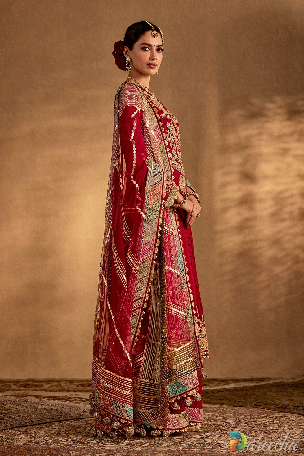 Model wearing an earthy maroon embroidered sharara suit with gold marodi ka kaam on the neckline and an organza dupatta.